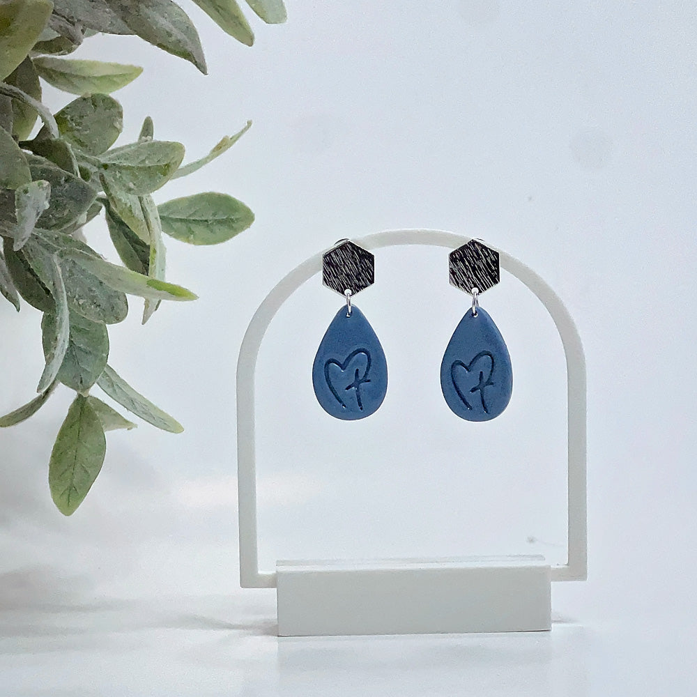Blue teardrop earrings with heart and cross design on a white stand 